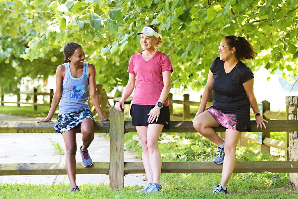 Three athletes in Blyss Running apparel pose by a fence in a park.